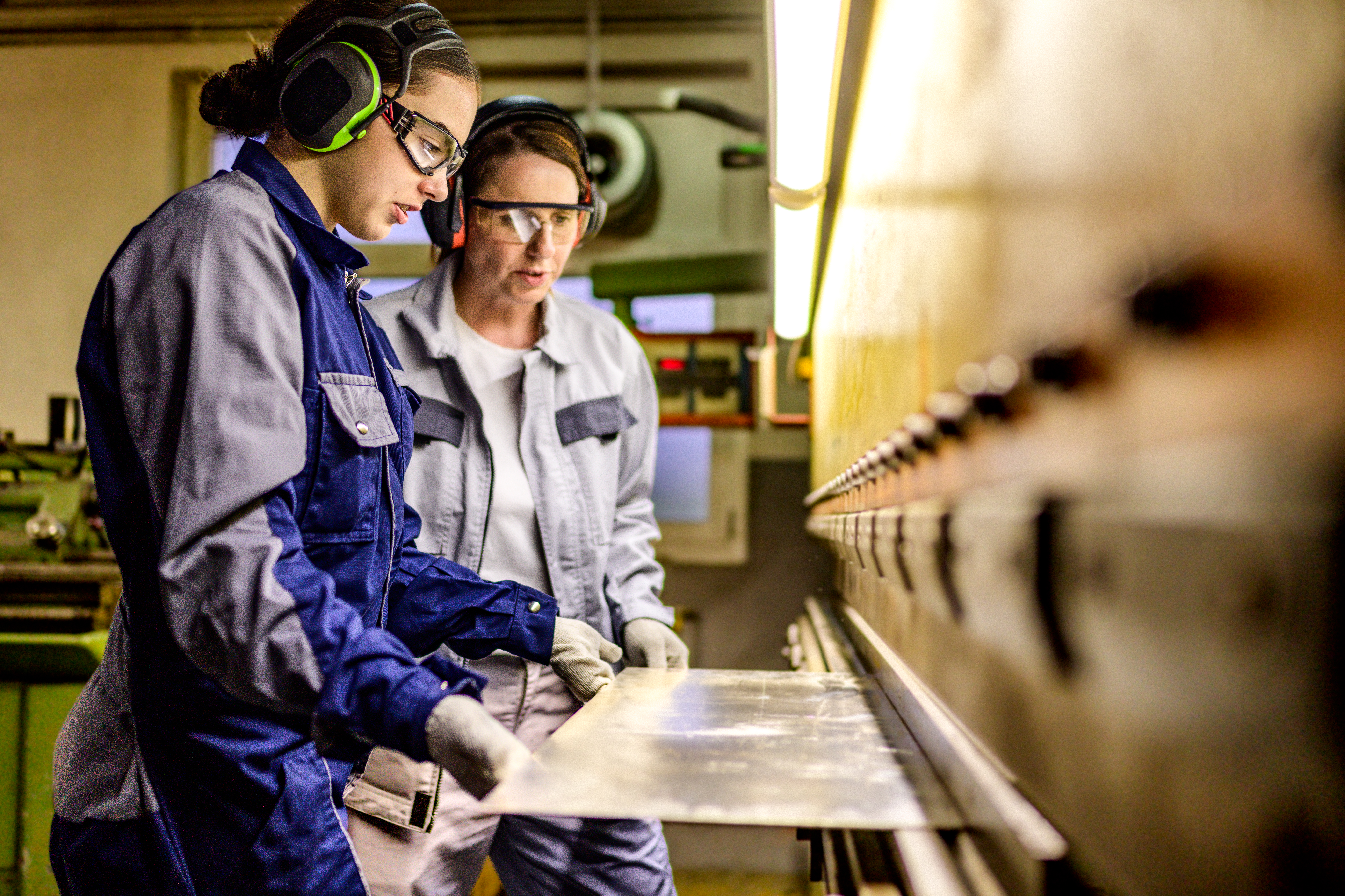 Two caucasian women wearing safety gear attentively manage a press brake machine, showcasing precision in a manufacturing setting.